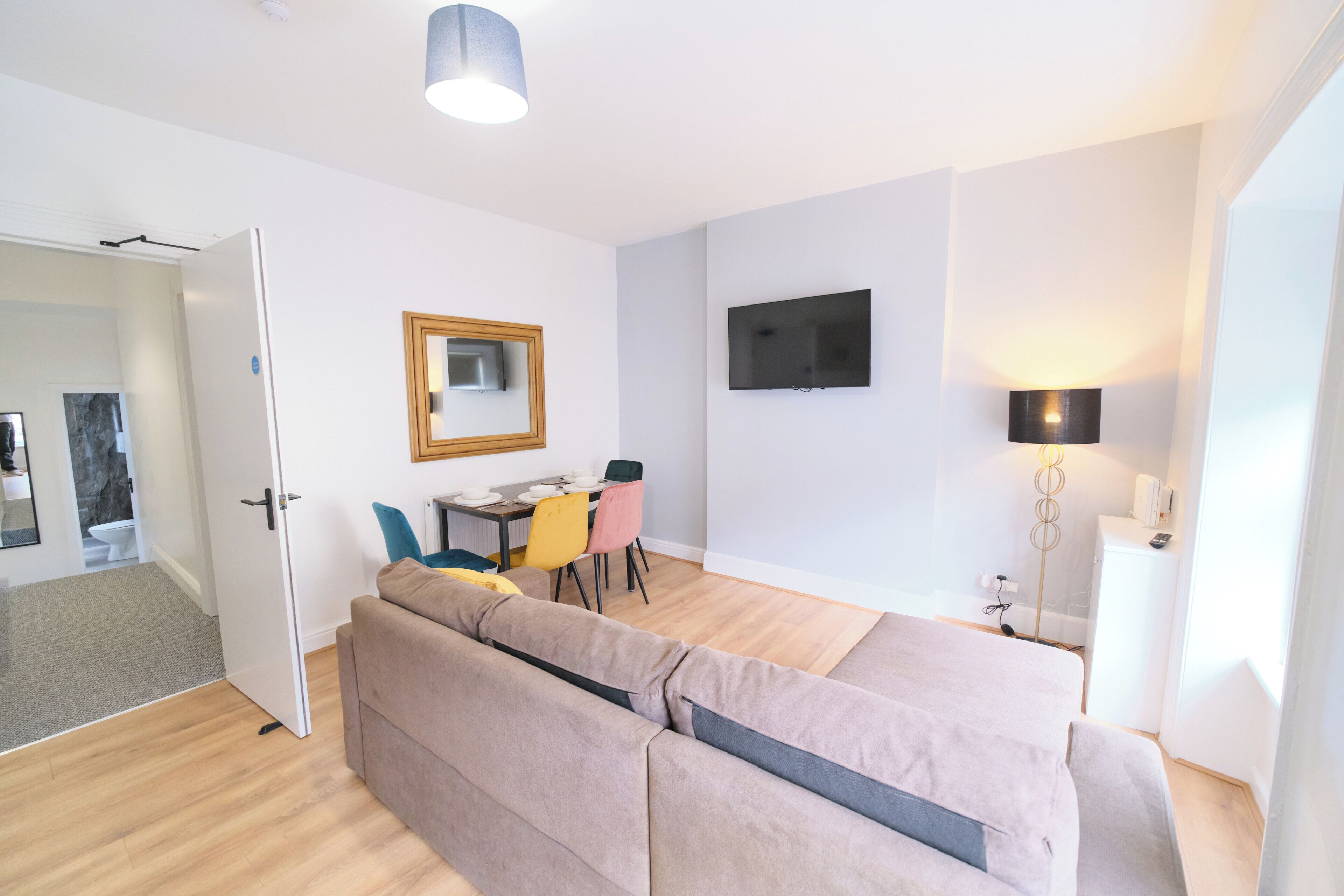 A modern, well-lit room with a sofa, a dining table with chairs, a flat-screen TV mounted on the wall, and a floor lamp.