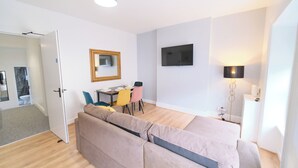 A modern, well-lit room with a sofa, a dining table with chairs, a flat-screen TV mounted on the wall, and a floor lamp. - Violet and Co (Derry/Londonderry)