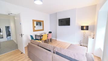 A modern, well-lit room with a sofa, a dining table with chairs, a flat-screen TV mounted on the wall, and a floor lamp.