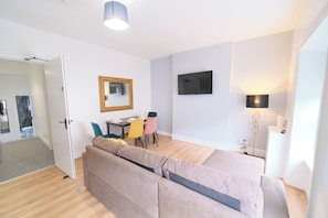 A modern, well-lit room with a sofa, a dining table with chairs, a flat-screen TV mounted on the wall, and a floor lamp. - Violet and Co (Derry/Londonderry)