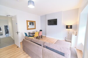 A modern, well-lit room with a sofa, a dining table with chairs, a flat-screen TV mounted on the wall, and a floor lamp.
