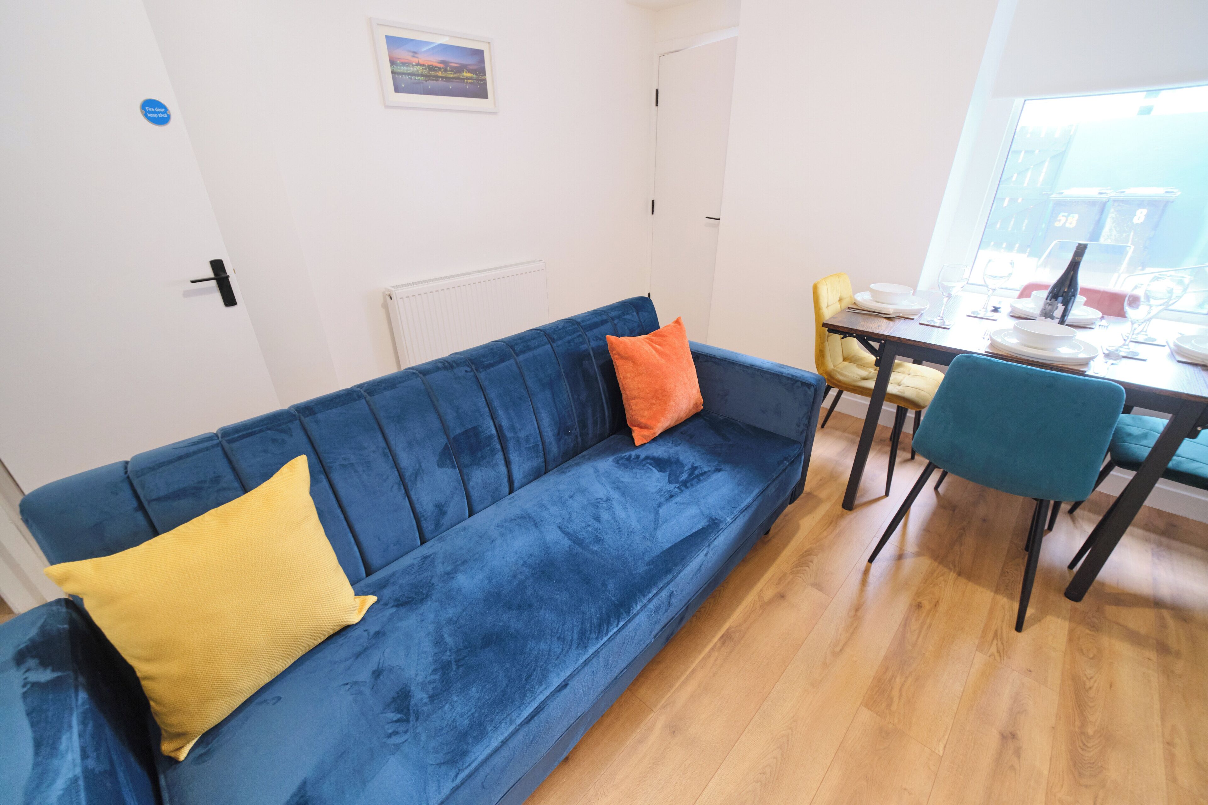 A blue sofa with yellow and orange cushions, a dining table with chairs, and a window with a view of buildings.