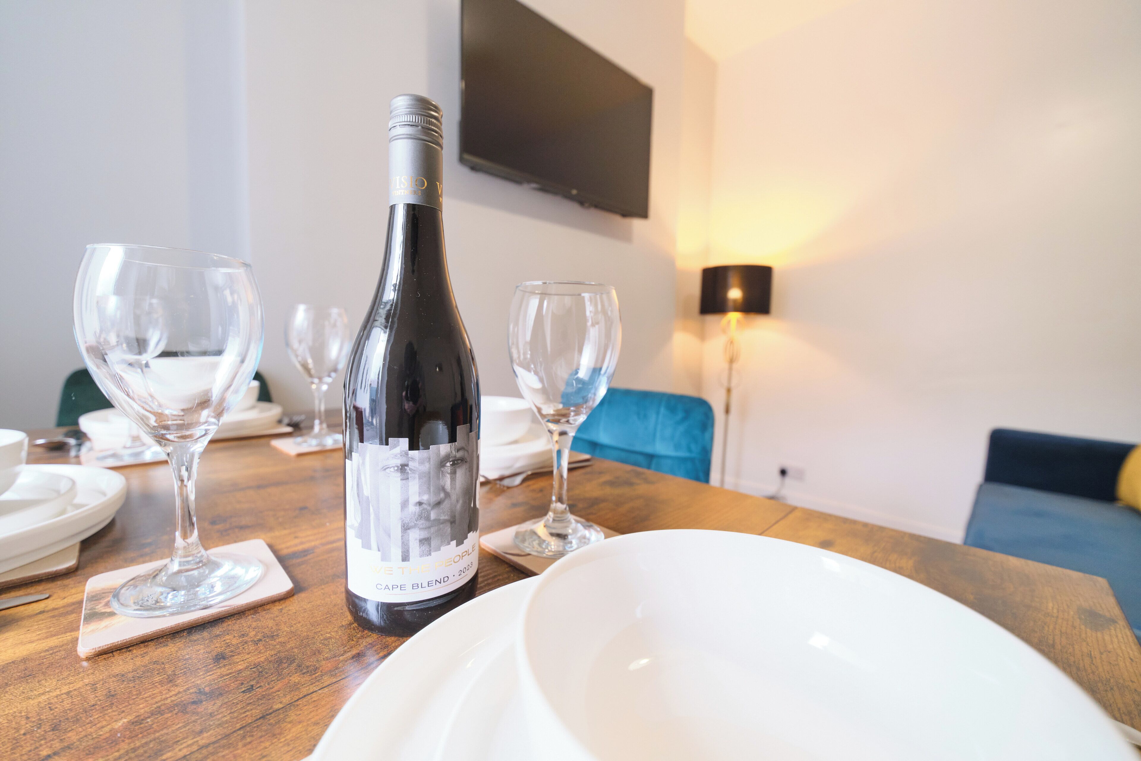 A dining table with a bottle of wine, wine glasses, and plates, set in a room with a sofa and a TV mounted on the wall.