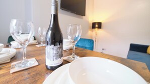 A dining table with a bottle of wine, wine glasses, and plates, set in a room with a sofa and a TV mounted on the wall. - Violet and Co (Derry/Londonderry)