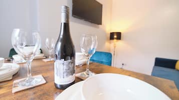 A dining table with a bottle of wine, wine glasses, and plates, set in a room with a sofa and a TV mounted on the wall.