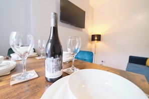 A dining table with a bottle of wine, wine glasses, and plates, set in a room with a sofa and a TV mounted on the wall. - Violet and Co (Derry/Londonderry)