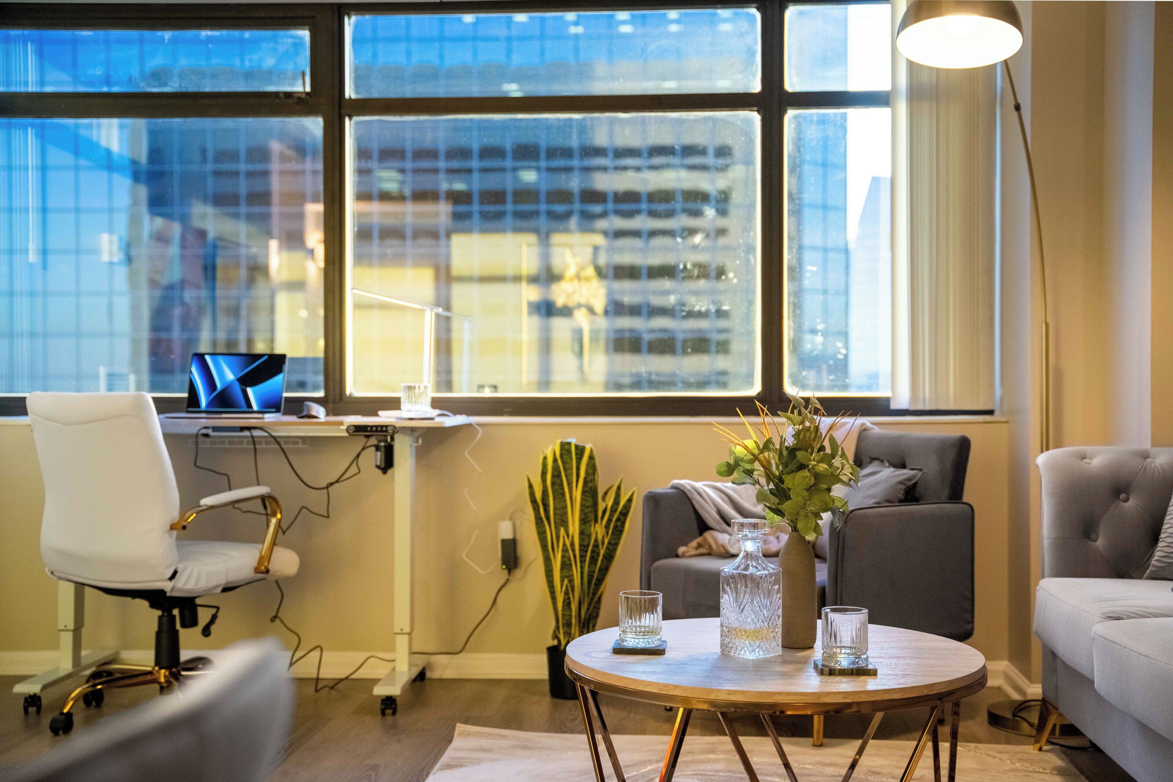 A modern office space with a desk, computer, and office chair, a round table with two glasses, and a sofa.