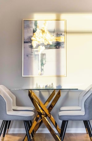 Een eettafel met een glazen blad en een gouden, kruisvormige voet, omringd door twee grijze fauteuils.