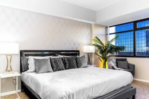 Een hotelkamer met een groot bed, een nachtkastje, een lamp, een potplant en uitzicht op de stad door een raam.