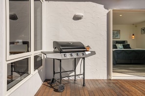 Un barbecue sur une terrasse en bois, avec un mur blanc à texture et une porte-fenêtre coulissante donnant sur une chambre.