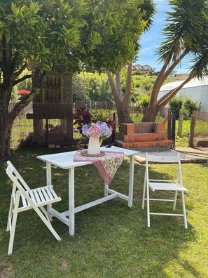 Ein weißer Tisch im Freien mit einer floralen Dekoration, zwei weiße Stühle und ein gemauerter Grill in einem grünen Garten mit Bäumen.