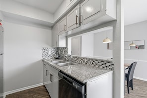 A modern kitchen with white cabinets, a granite countertop, a dishwasher, and a tiled backsplash. - Chic 2BR Oasis in Houston (Houston)