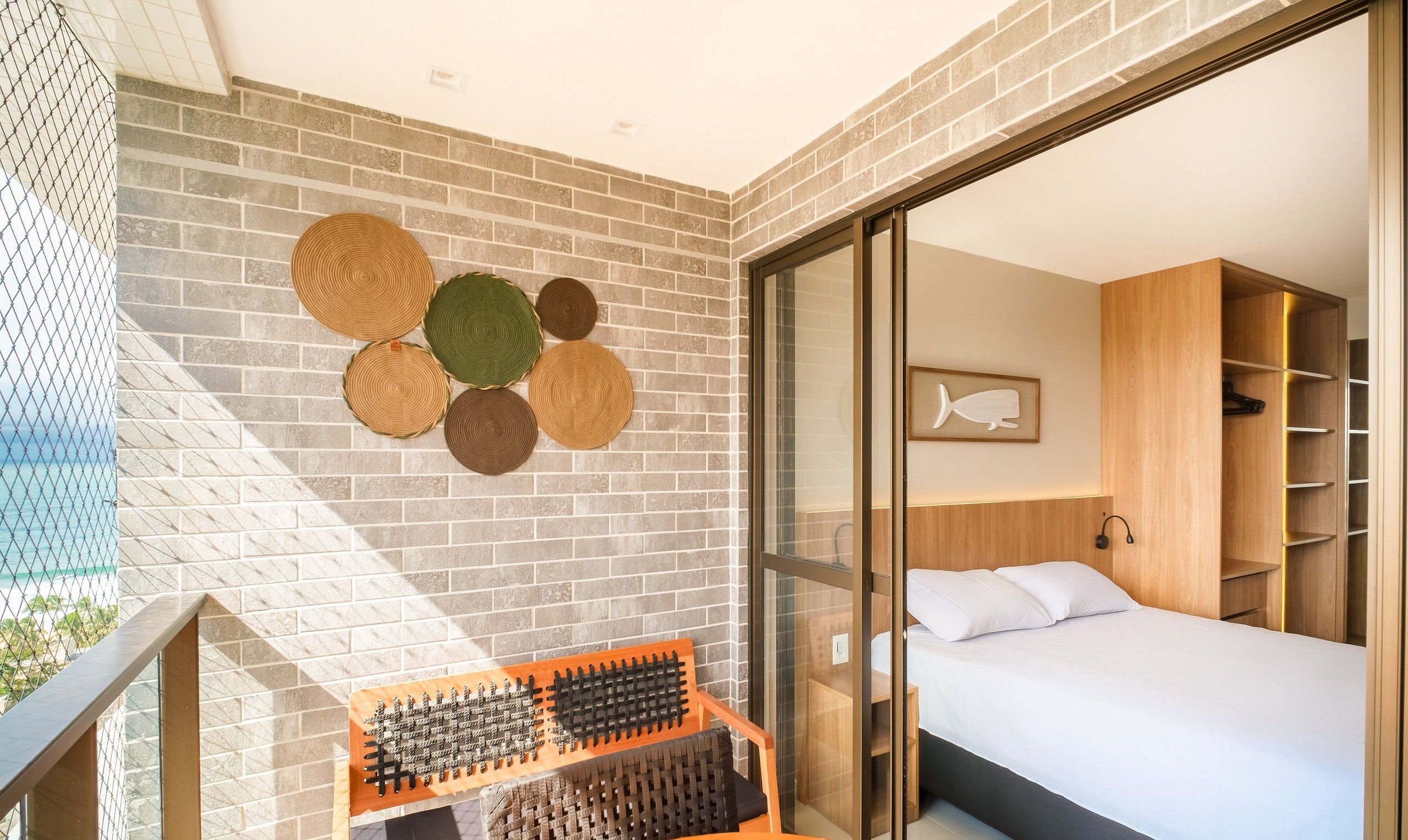 A modern hotel room with a brick wall, a bed with white linens, a wooden wardrobe, and a wall-mounted shelf with woven mats.