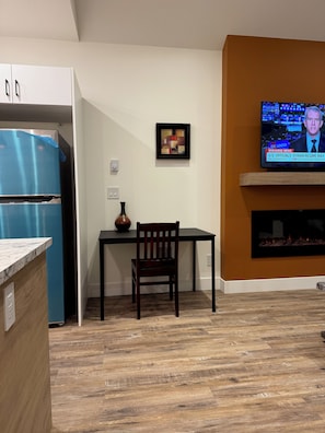 A modern kitchen with a blue refrigerator, a dining table with a chair, a framed abstract painting, and a television displaying news.