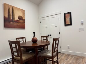 A dining area with a round table, four chairs, a vase, a painting, and a door with a light switch.