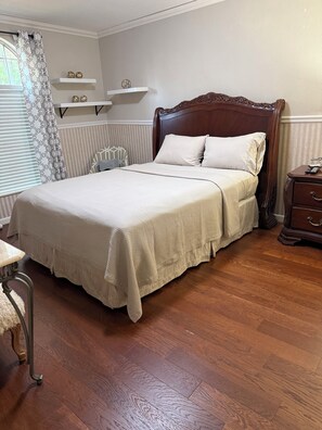 Een netjes opgemaakt bed met een houten hoofdbord, een nachtkastje met een lamp en een stoel met een kussen.