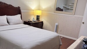 Quarto de hotel com uma cama grande, mesa de cabeceira com abajur, pequena mesa com cafeteira e radiador.