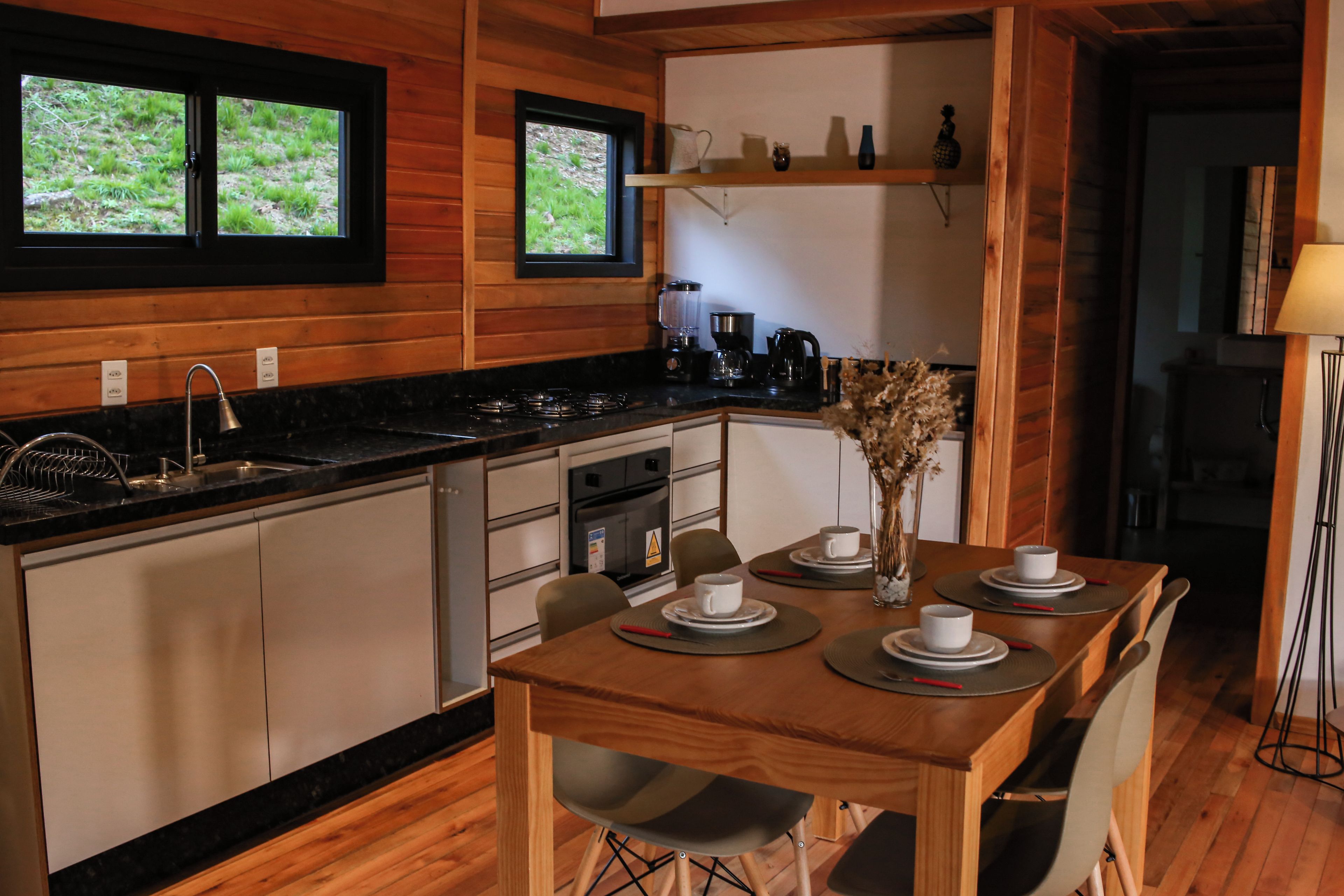 A modern kitchen with a wooden interior, a dining area with a table set for four, and a view of greenery through the windows.