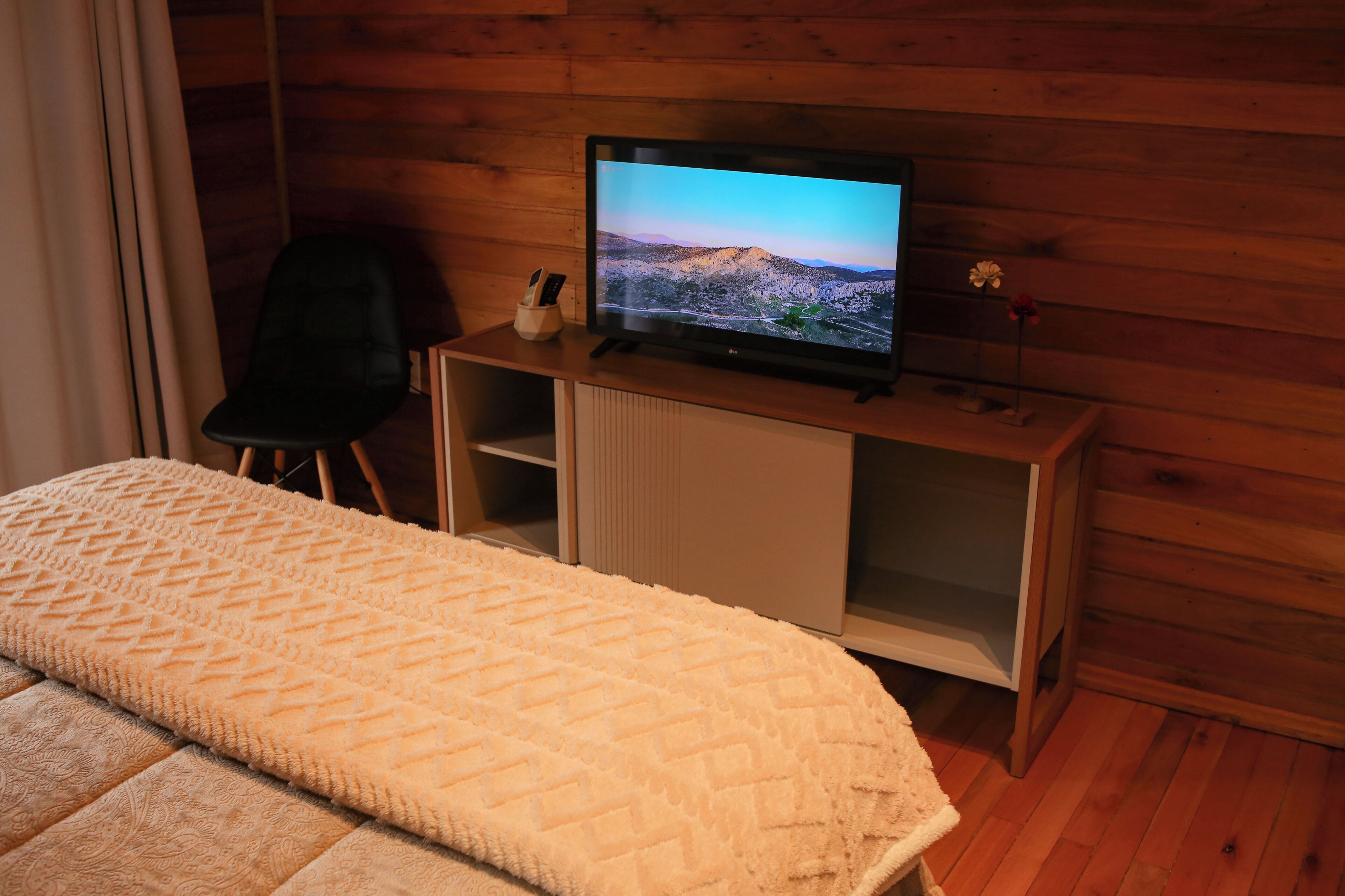 Un dormitorio con una cama, televisión sobre un mueble de madera, una silla y una pequeña mesa con un florero.