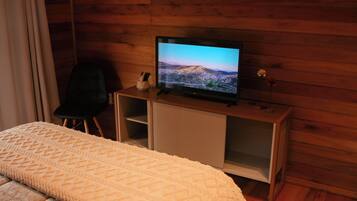 Un dormitorio con una cama, televisión sobre un mueble de madera, una silla y una pequeña mesa con un florero.