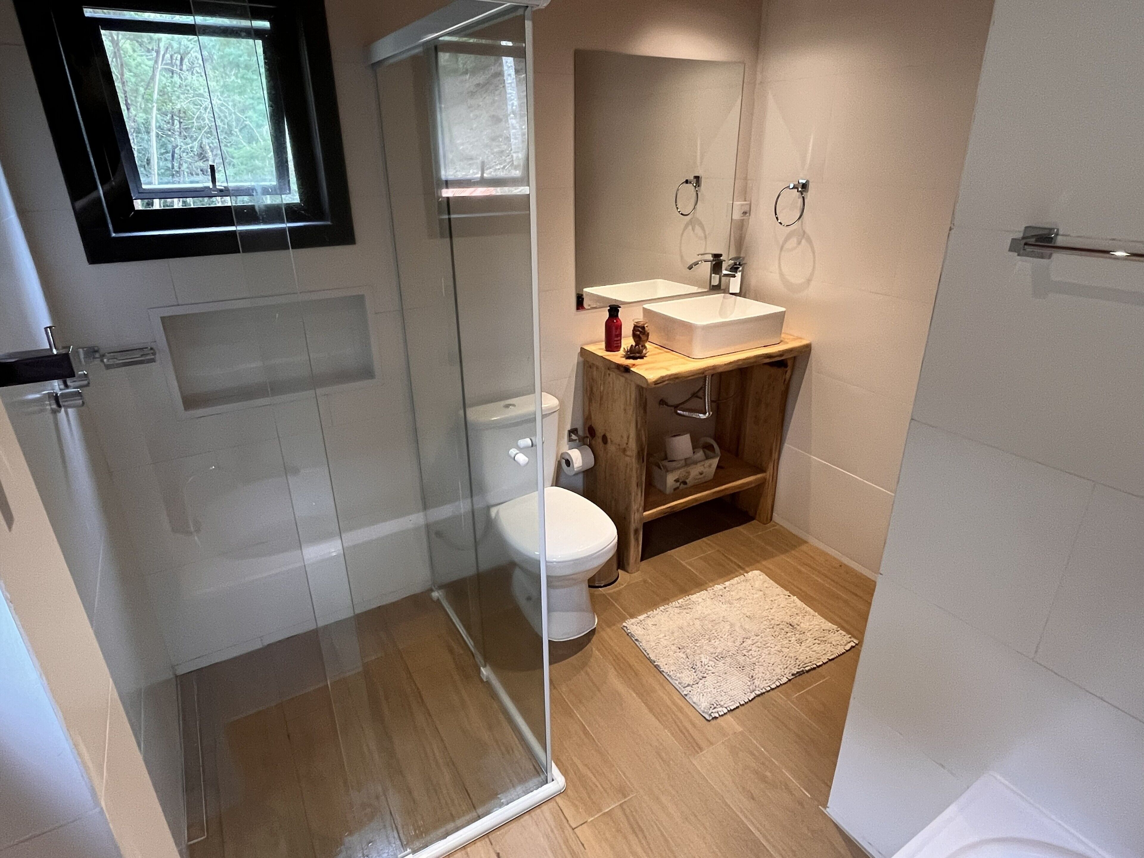 A modern bathroom with a glass shower enclosure, a white toilet, a wooden vanity with a sink, and a small rug on the floor.