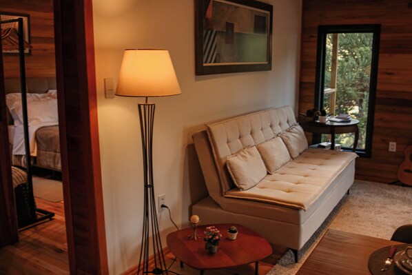 A cozy room with a sofa, a floor lamp, a small table with a vase, and a window with a view of greenery.