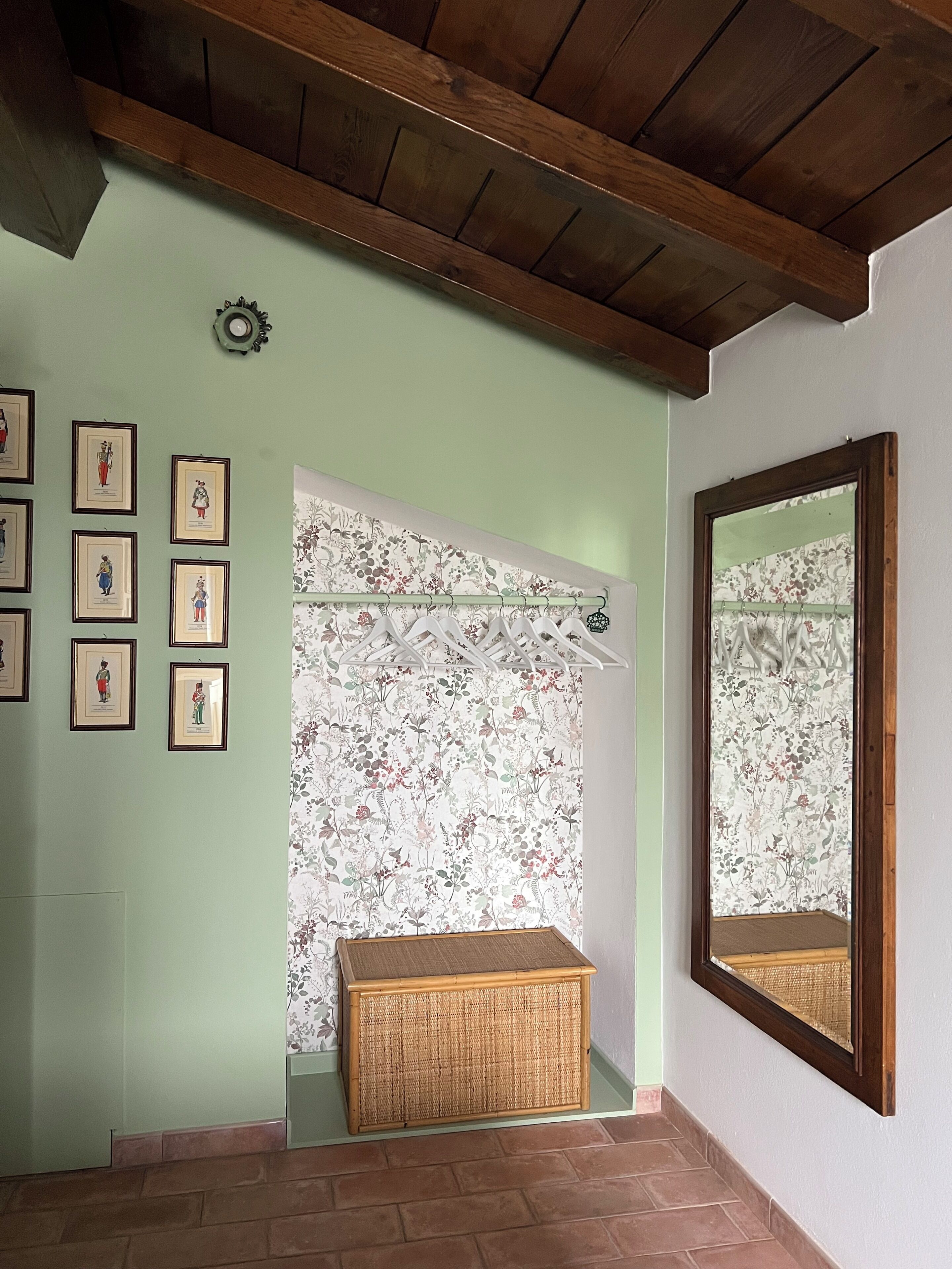 Una stanza con soffitto in legno, uno specchio fissato al muro, un cesto in vimini e una tenda con motivi floreali.