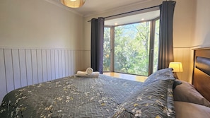 Un dormitorio con una cama grande, vistas a árboles por una ventana y una mesita de noche con una lámpara.