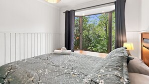 Quarto com uma janela grande, uma cama com colcha estampada e uma mesa de cabeceira com abajur.