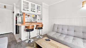 Un espacio habitable funcional con kitchenette, un sofá gris y dos taburetes de bar.