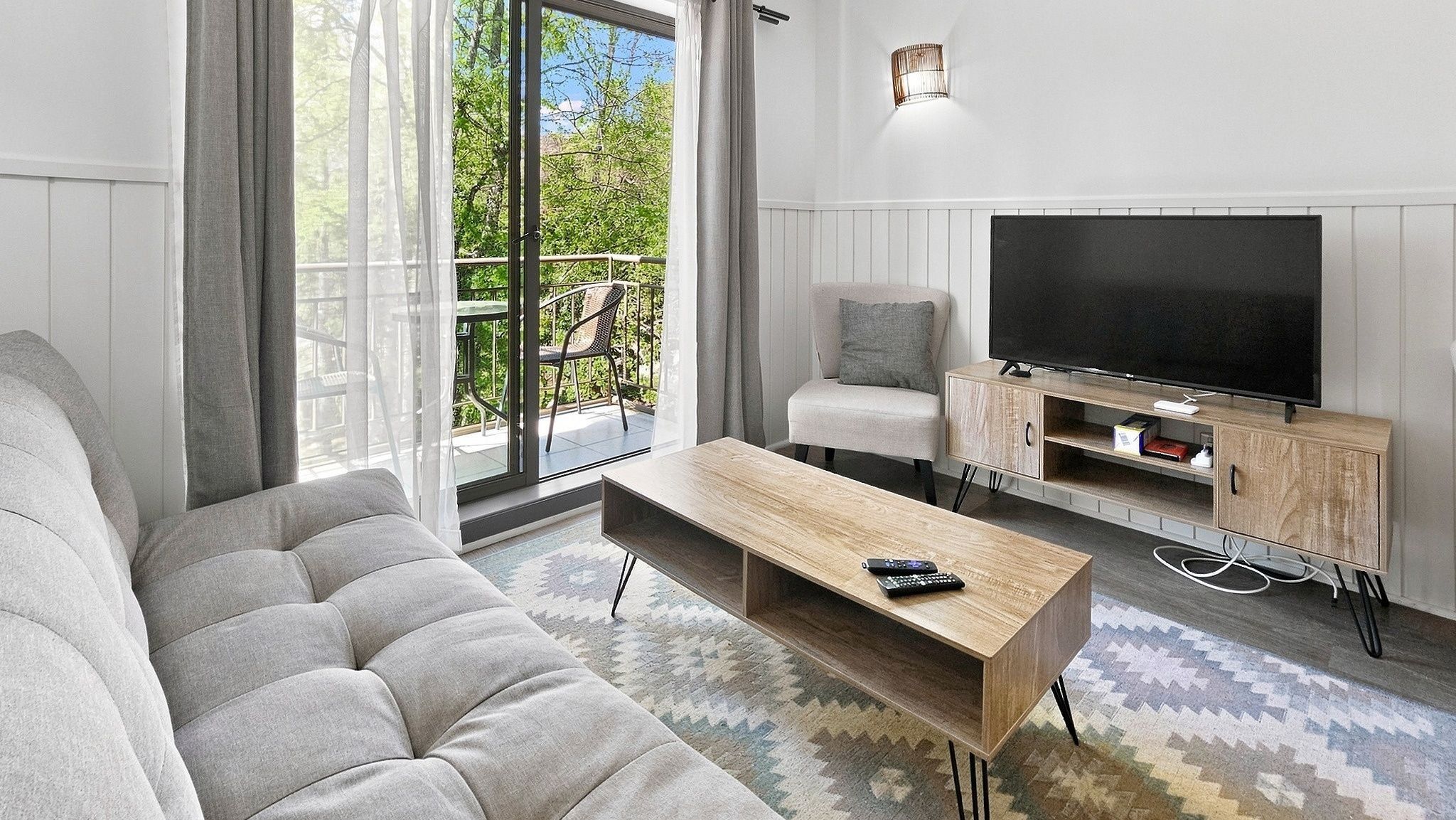 A modern living room with a sofa, a coffee table, a TV on a wooden stand, and a balcony with outdoor furniture.