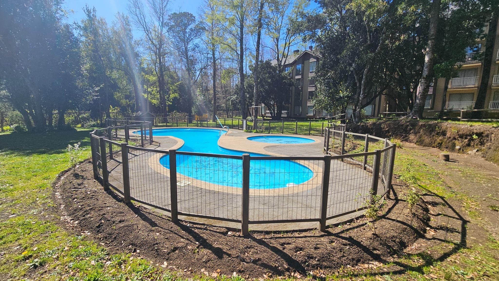 Une piscine circulaire entourée d’une clôture, bordée d’arbres et avec un bâtiment visible au loin.