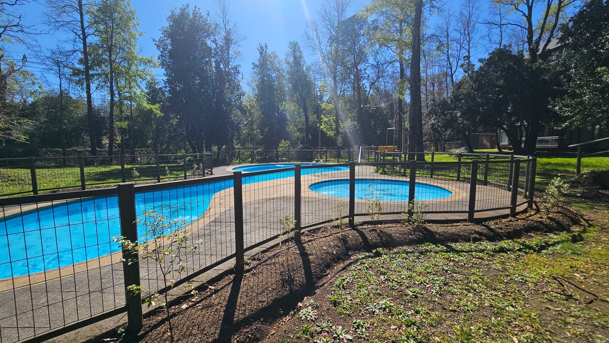 A fenced outdoor swimming pool with a circular design, surrounded by trees and a grassy area.
