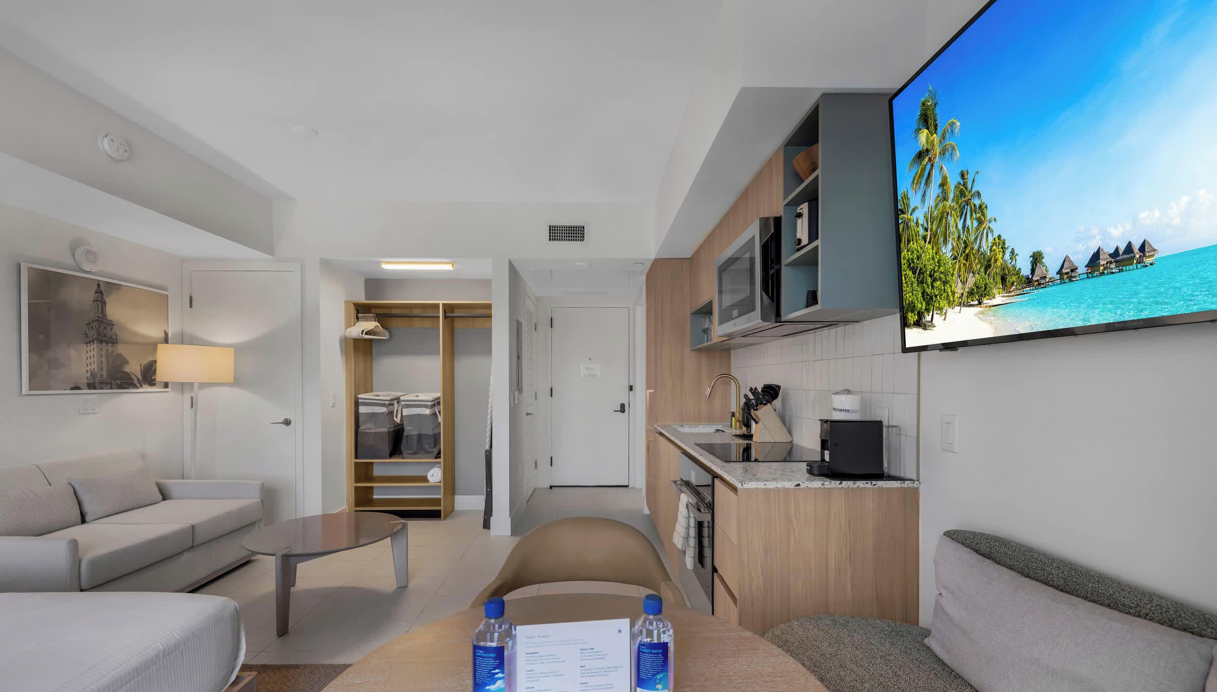 A modern hotel room with a living area, kitchenette, and a large TV screen displaying a tropical beach scene.