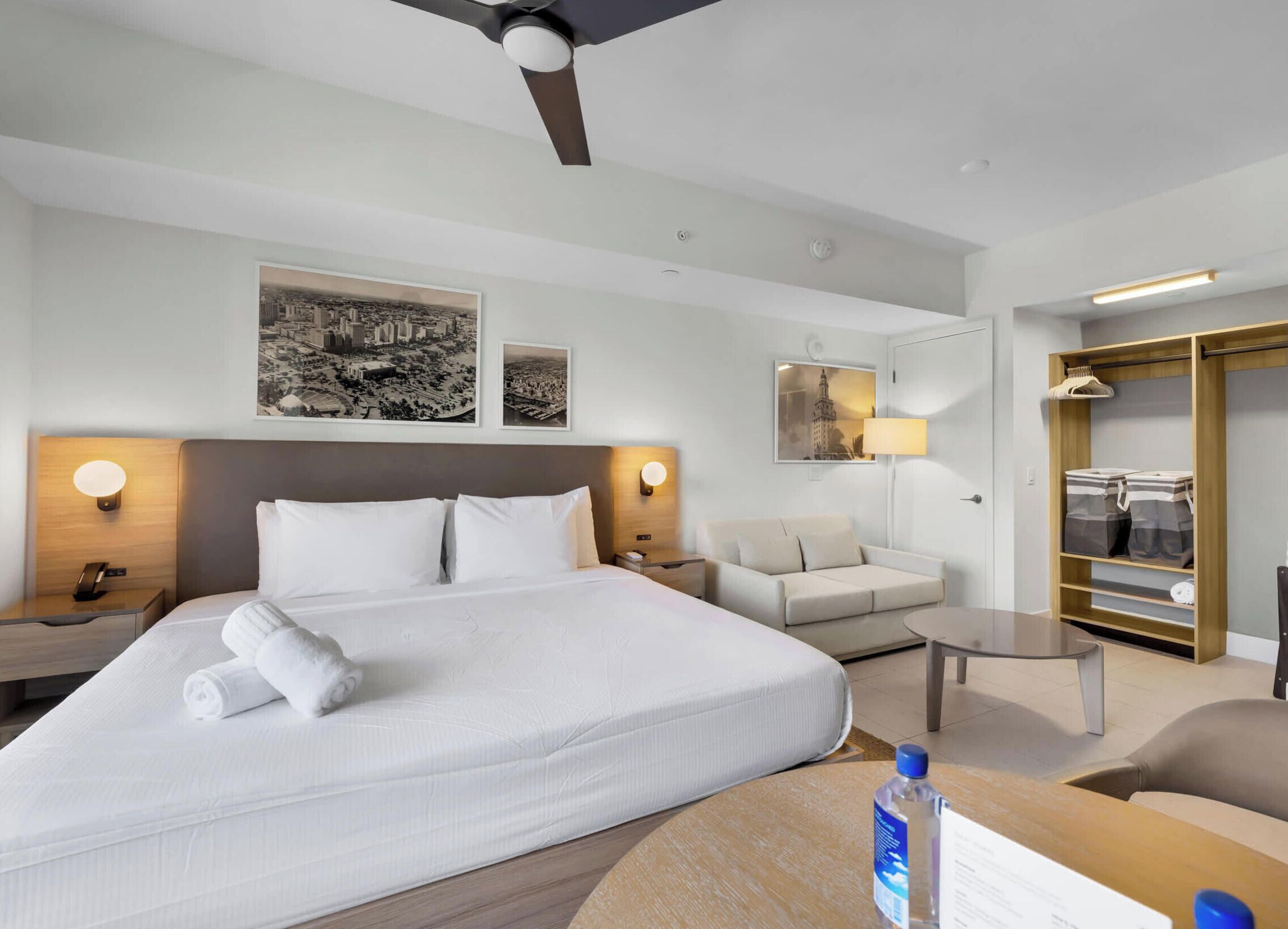 A hotel room with a large bed, a bedside table, a nightstand, a desk, a chair, a sofa, a wardrobe, and a ceiling fan.