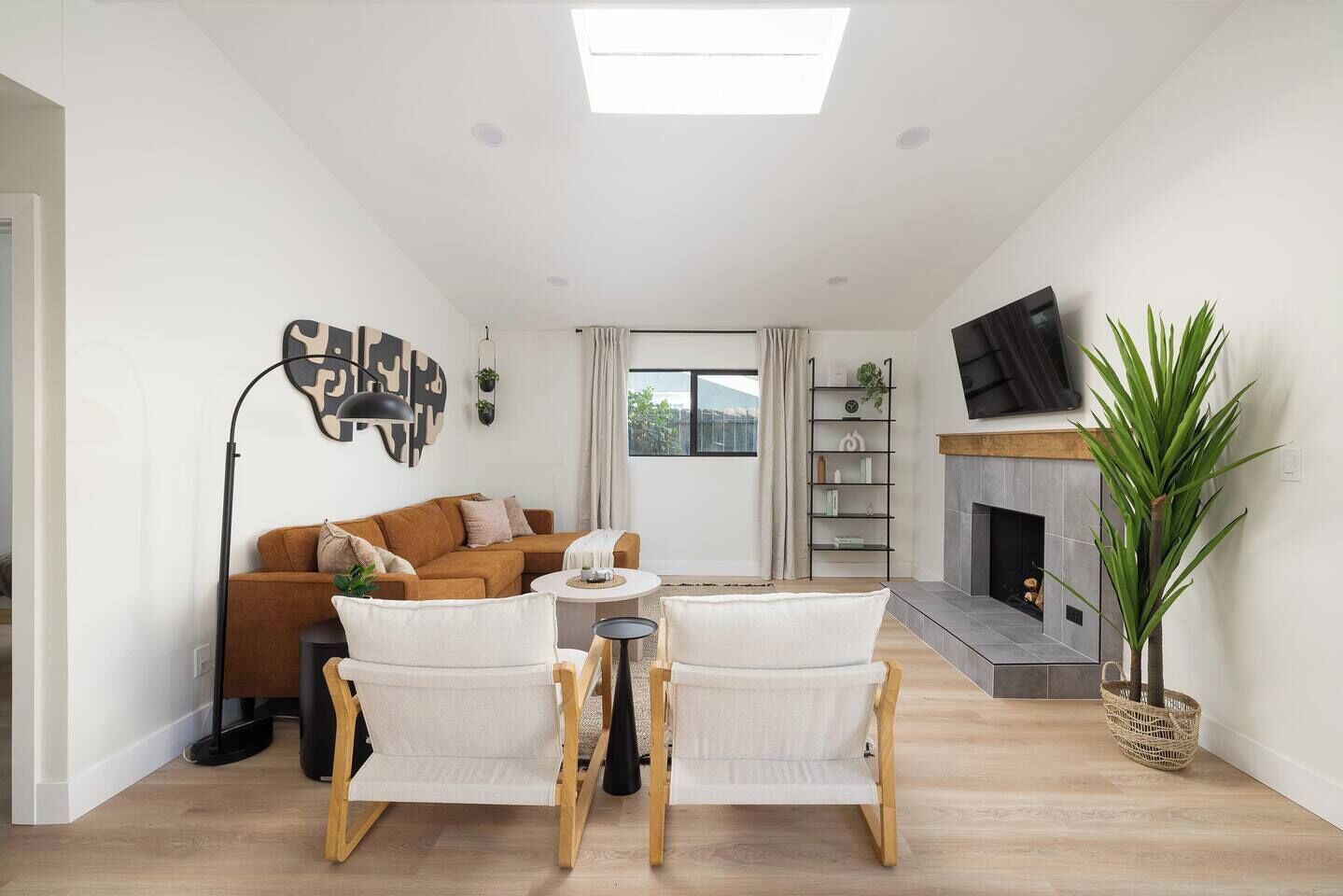 A living room with a sofa, two armchairs, a coffee table, a fireplace, a TV mounted on the wall, and a window with curtains.