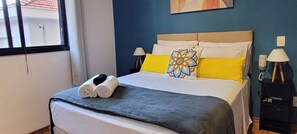 Ein ordentlich bezogenes Bett mit blauer Decke, zwei Kissen und einem Zierkissen. Auf jeder Seite des Bettes steht eine Nachttischlampe.