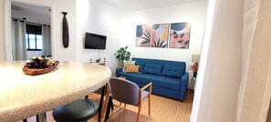 Ein modernes Wohnzimmer mit einem blauen Sofa, einem wandmontierten Flachbildfernseher und einem Essbereich mit rundem Tisch und Stühlen.