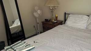 A neatly made bed with white bedding, a bedside lamp, a fan, and a mirror on the wall.