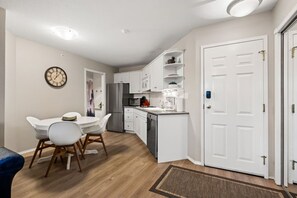 A compact kitchen with white cabinets, a stainless steel refrigerator, and a microwave. There is a dining area with a white table and chairs. - Riviera | Balcony w/ Mountain Views | Pets | Golf (Fairmont Hot Springs)