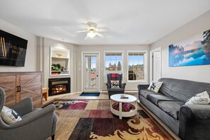 A living room with a fireplace, a sofa, a coffee table, and a large window with a view. - Riviera | Balcony w/ Mountain Views | Pets | Golf (Fairmont Hot Springs)