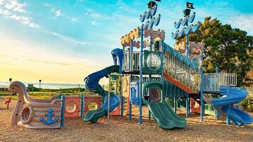 A playground with slides and climbing structures, set against a backdrop of trees and a clear sky.