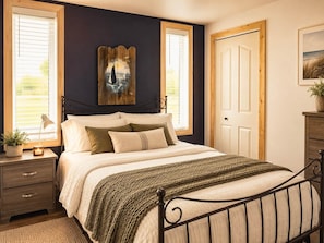 A bedroom with a bed, bedside tables, a nightstand, a window with blinds, a door, and a wall-mounted artwork.
