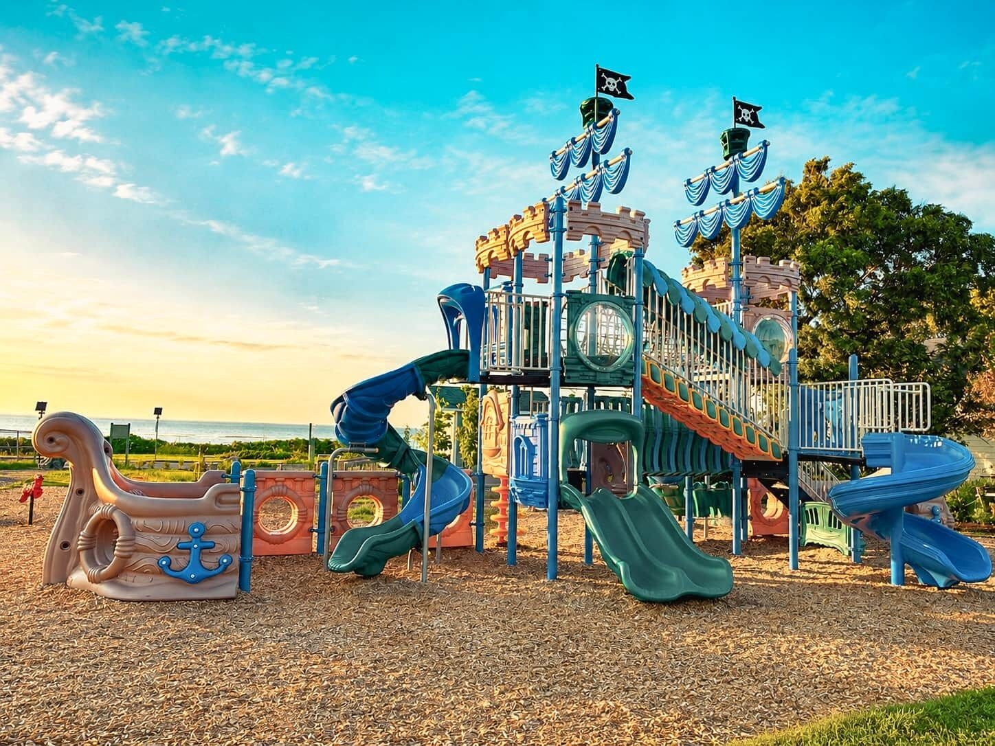Un parque infantil con toboganes y estructuras para escalar, frente a un fondo de árboles y un cielo despejado.