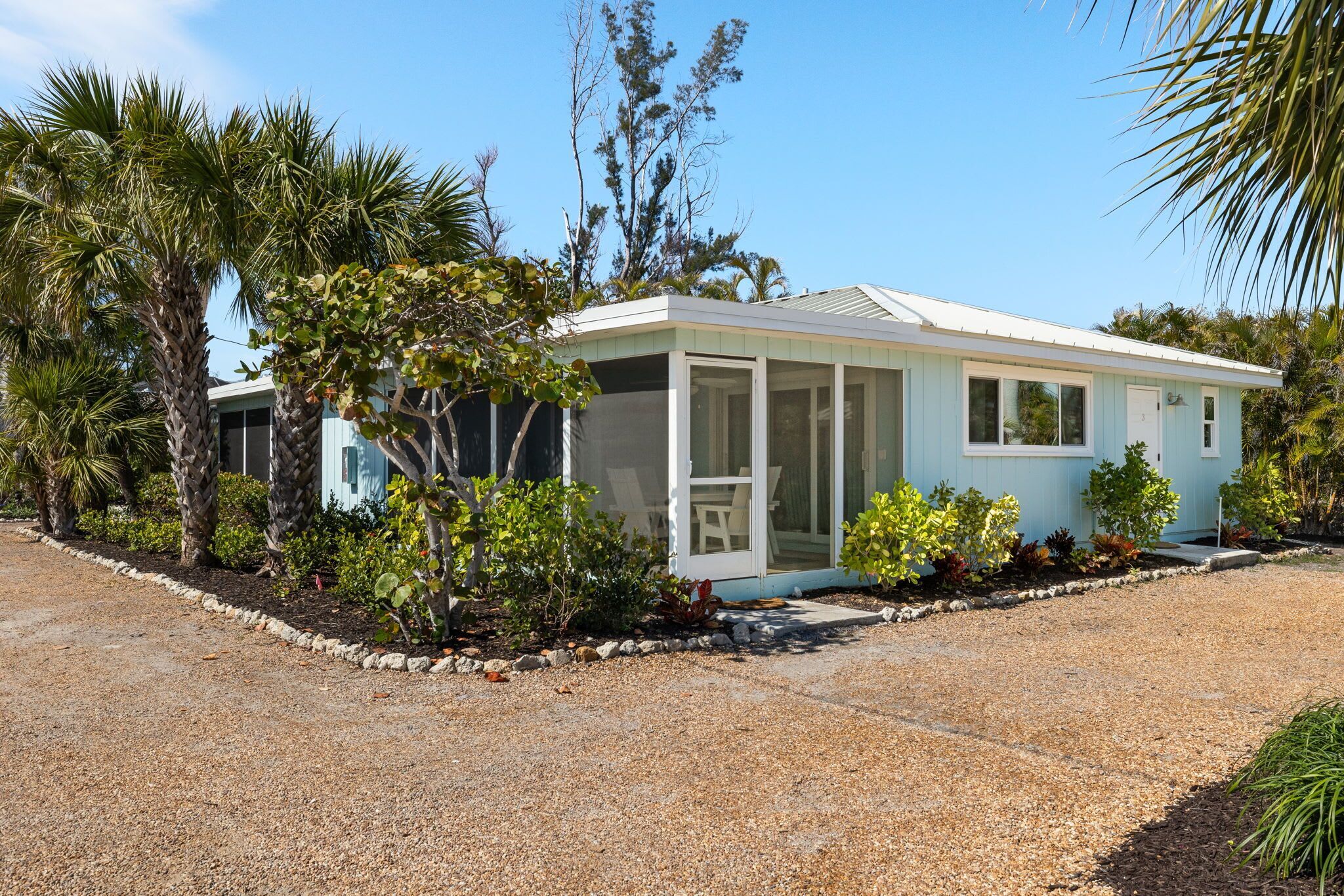 A single-story house with a covered porch, surrounded by a gravel driveway and various tropical plants.