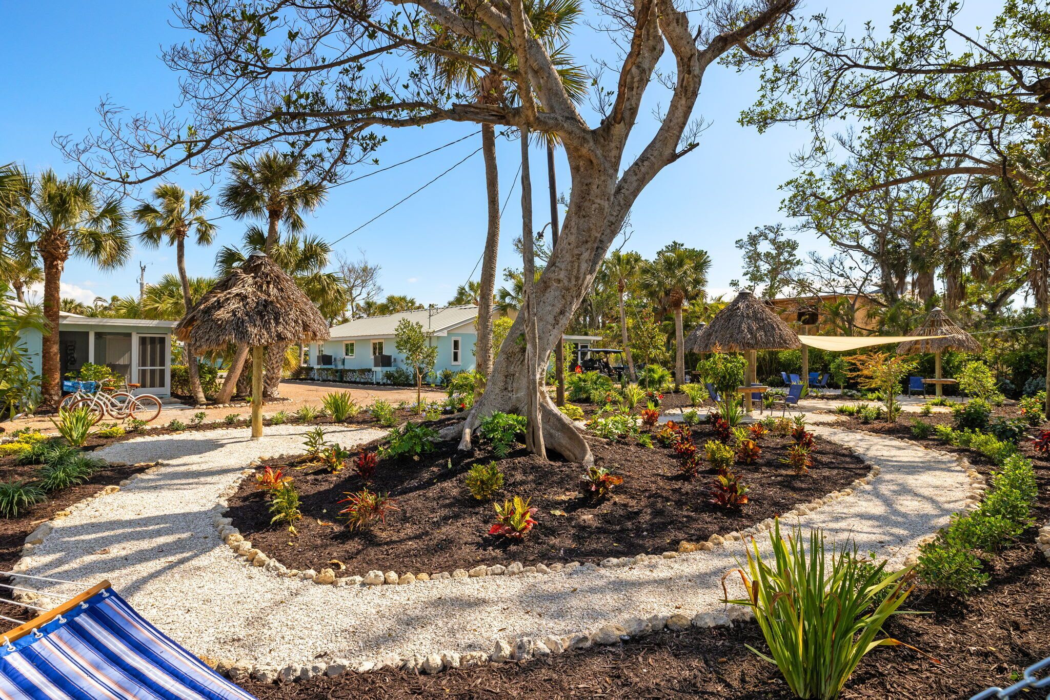 A garden with a tree, palm trees, and a building with a thatched roof.