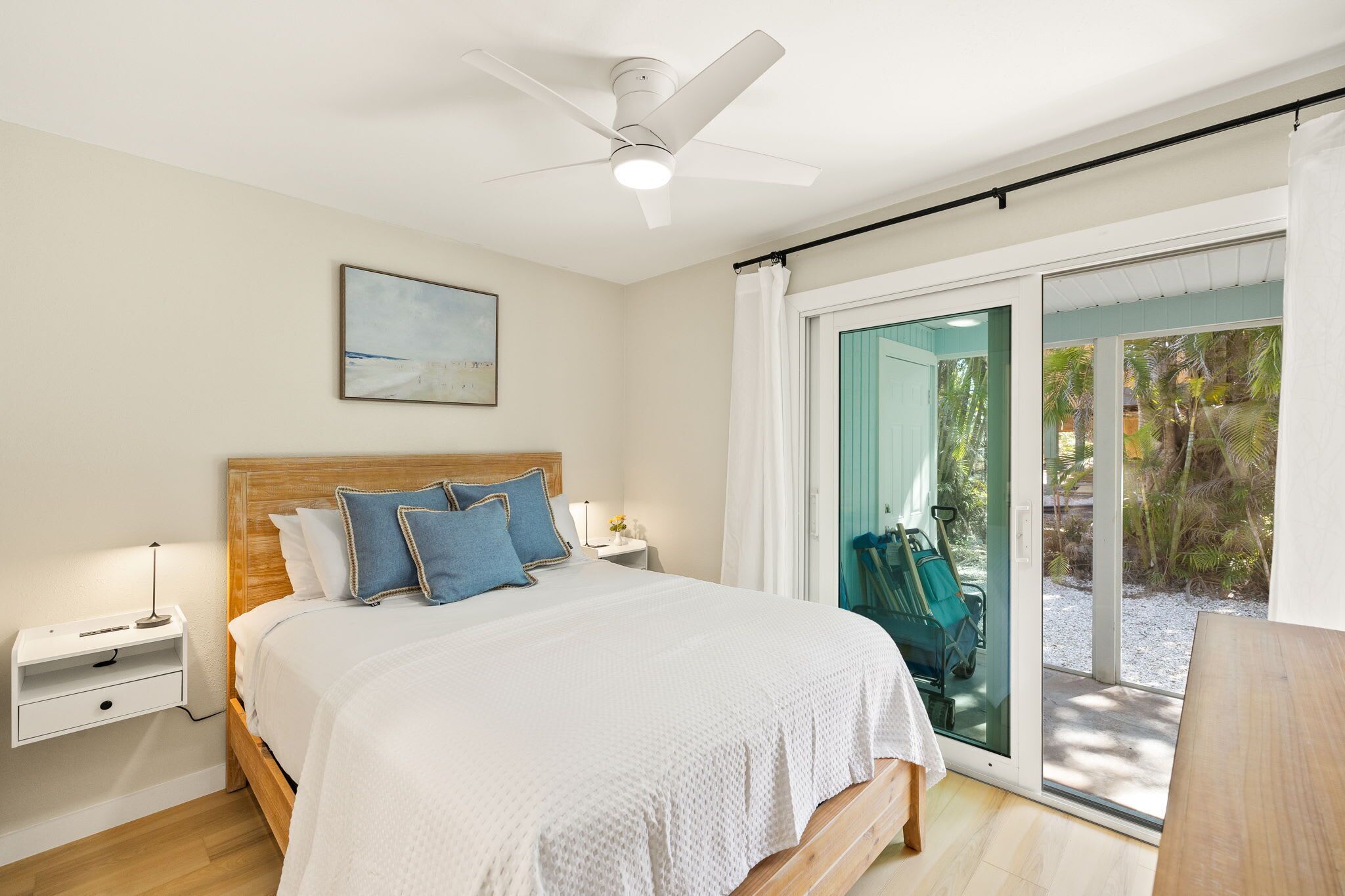 Een slaapkamer met een bed, nachtkastjes, een plafondventilator, een raam met uitzicht op palmbomen en een schuifdeur met glas.