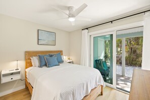 A bedroom with a bed, nightstands, a ceiling fan, a window with a view of palm trees, and a sliding glass door.