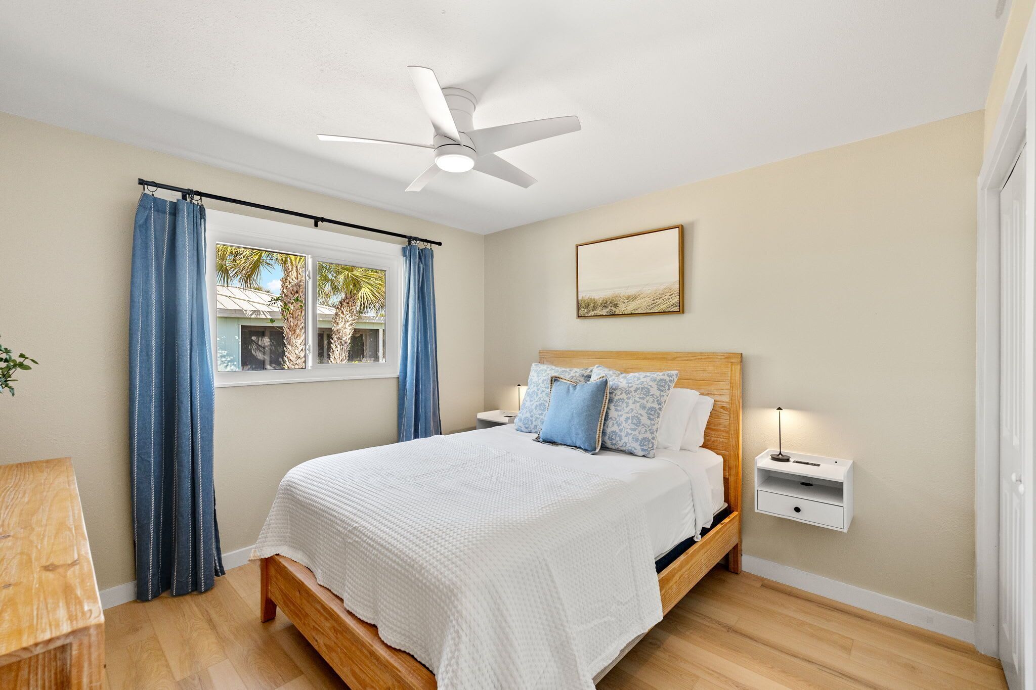 A bedroom with a bed, a ceiling fan, a nightstand with a lamp, a window with curtains, and a wall-mounted picture.