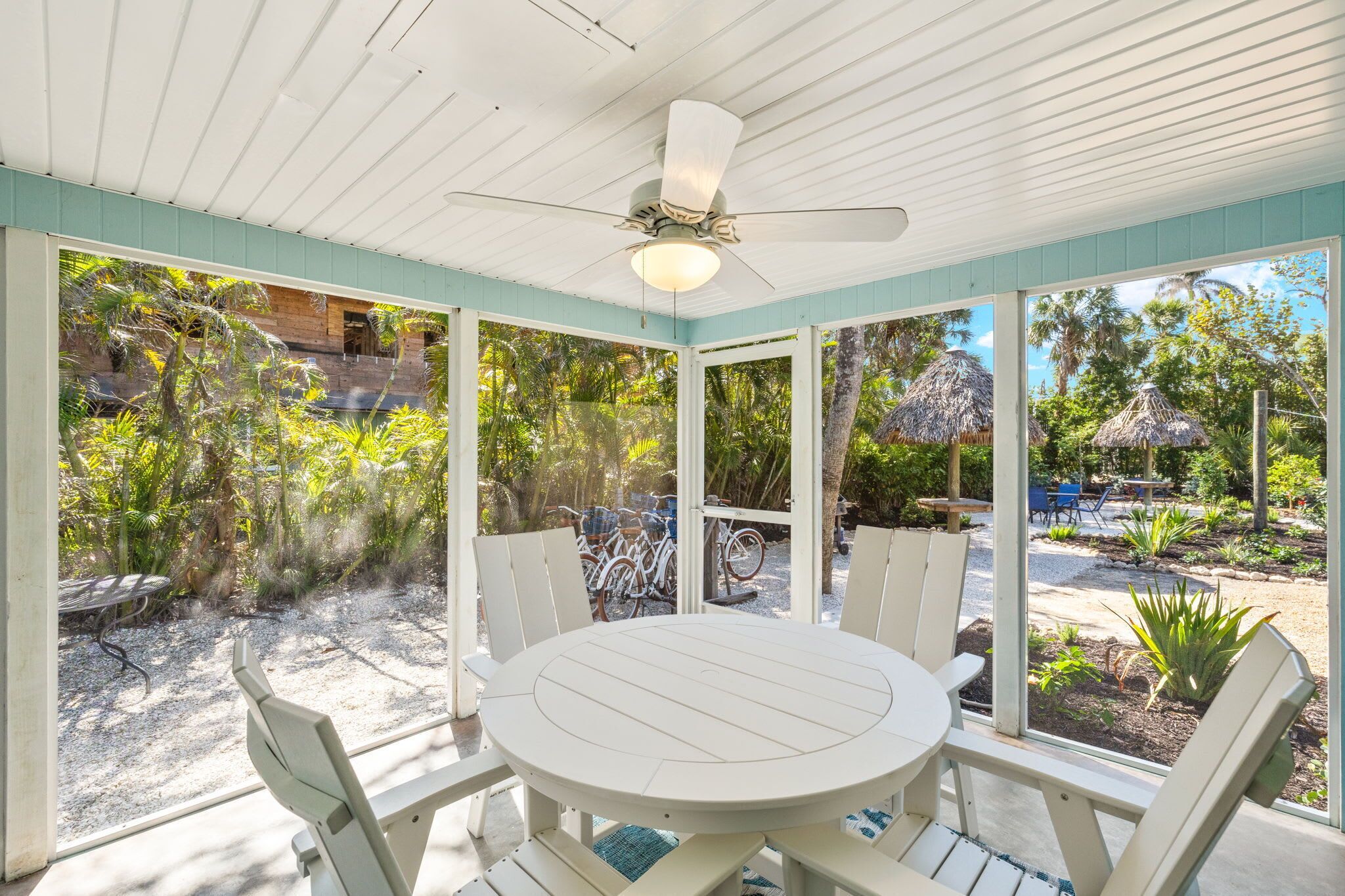A covered patio with a round table and chairs, a ceiling fan, and a view of a tropical garden.